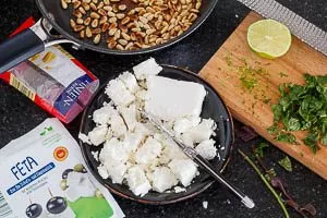 Feta zerkleinern und Minze und Limette vorbereiten. Pinienkerne in der Pfanne anbraten.