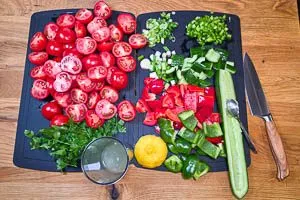 Rote Paprika, Frühlingszwiebeln, Salatgurke, Tomaten und Knoblauchzehen vorbereiten und in kleine Stücke schneiden. Petersilie waschen und Stängel entfernen, Zitrone auspressen.