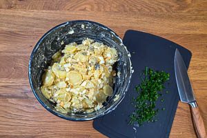Schnittlauch kleinschneiden und über den Kartoffelsalat mit Cornichons, Zwiebeln und Eiern geben