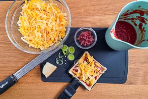 Teig auf der oberen Platte vorbacken, Tomatensoße, Parmesan, Lauch, Rohschinkenwürfel und geriebenen Käse darauf verteilen.