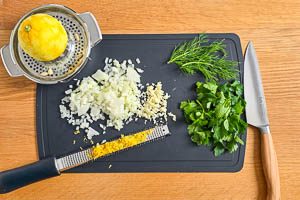 Zwiebeln und Knoblauch w&uuml;rfeln, Petersilie und Dill waschen, trocknen und Bl&auml;tter abzupfen, Zitronenzeste abreiben, Zitrone auspressen