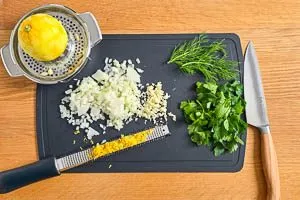 Zwiebeln und Knoblauch w&uuml;rfeln, Petersilie und Dill waschen, trocknen und Bl&auml;tter abzupfen, Zitronenzeste abreiben, Zitrone auspressen