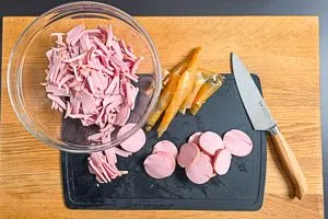 Fleischwurst pellen, in Scheiben und dann in Streifen schneiden