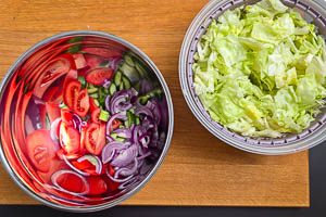 Tomaten, Gurken und Zwiebeln in eine Sch&uuml;ssel geben. Eisbergsalat daraufgeben.
