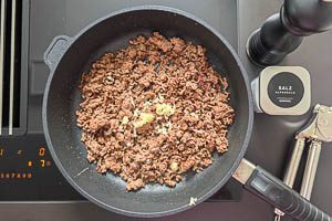 Rinderhack in einer Pfanne mit neutralem Pflanzen&ouml;l, Salz, schwarzem Pfeffer und einer gepressten Knoblauchzehe anbraten.